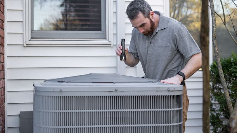 HVAC technician with a flashlight inspecting an outdoor AC condenser beside a house